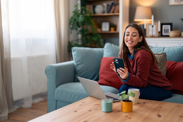 Cheerful lady multitasks with a laptop and phone, smartly dressed in casual attire, while nestled on a cozy sofa within a well-adorned living space.