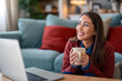 © Dorde - A cheerful Caucasian woman participates in an online chat, cradling a mug, dressed casually in her cozy living room, using a laptop placed on a table.