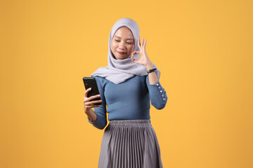 happy asian indonesian muslim woman holding smart phone giving ok finger gesture on isolated yellow background