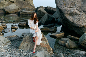 Poster - Serene woman in white dress with red shoes sitting on rocky shoreline by the ocean