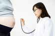 © TAGSTOCK2 - 妊婦の大きなお腹に聴診器をあて診察をしようとする若い女医の横姿