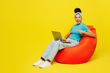 Wall Mural - Full body young Latin IT woman wears blue top t-shirt headband casual clothes sit in bag chair use work point index finger on laptop pc computer isolated on plain yellow background. Lifestyle concept.