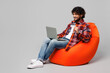 © ViDi Studio - Full body young IT Indian man wears red shirt casual clothes sit in bag chair using laptop pc computer hold credit bank card shopping online isolated on plain grey color background. Lifestyle concept.