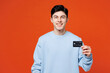 © ViDi Studio - Young smiling happy man he wears blue sweatshirt casual clothes hold in hand mock up of credit bank card show thumb up isolated on plain red orange color background studio portrait. Lifestyle concept.