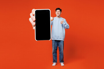 Wall Mural - Full body young man he wears blue sweatshirt casual clothes hold in hand use mobile cell phone with blank screen workspace area isolated on plain red orange color background studio. Lifestyle concept.