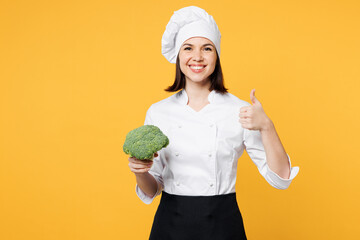 Wall Mural - Young fun chef cook baker woman wear white shirt black apron uniform toque chefs hat hold in hand green broccoli looking camera show thumb up isolated on plain yellow background. Cooking food concept.