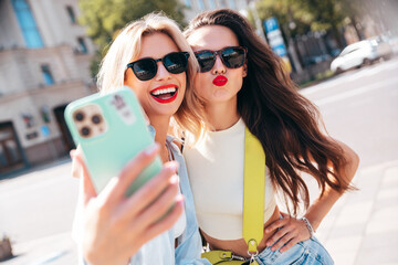 Wall Mural - Two young beautiful smiling female in trendy summer clothes. Sexy carefree women posing in the street. Positive models having fun, hugging in sunny day. With bright makeup, sunglasses, take selfie