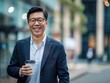 © hassan - Asian businessman with glasses, smiling with coffee cup, walking outside office building, street view.