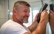 © Антон Сальников - A middle-aged man looks into the mirror and shaves the beard or hair with the electric shaver or trimmer at home
