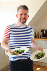 Adult male man smiling holding two plates of healthy food in kitchen, looking at camera, domestic life, real people, UK