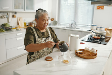 Senior woman standing in the kitchen and putting tea leaves in teapot to make black tea