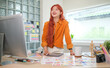 © wattana - Young female graphic designer standing at her desk surrounded by colour swatches and design tools.