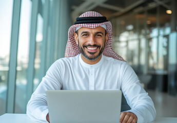 Smiling man in traditional attire working on a laptop in a modern office with large windows. The scene blends cultural heritage with professionalism.