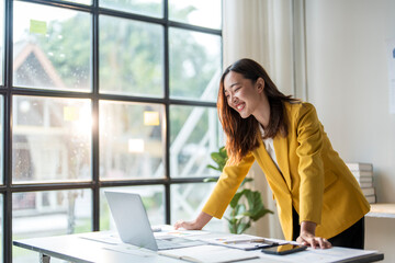 Wall Mural - young asian businesswoman working and smiling at laptop in sunny office