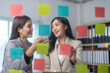 © Wasana - Two businesswomen smiling and brainstorming using sticky notes on glass wall in office meeting