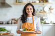 © Niks Ads - a beautiful smiling young indian woman in an apron holding a plate of food standing in a kitchen at home.