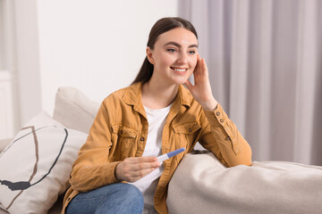 Wall Mural - Happy woman holding pregnancy test on sofa indoors