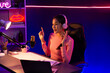 © Summit Art Creations - Smiling creative young beautiful Asian influencer talking in channel with positive podcast speech at night time, using laptop for presentation online at pink neon light modern studio. Stratagem.