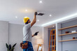© tynza - Electrician installing a new light fixture on the ceiling, with a toolbox and ladder in a modern living room.