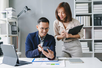 Two asian business people smiling and working together in a modern office, the woman holding a clipboard and the man using a smartphone