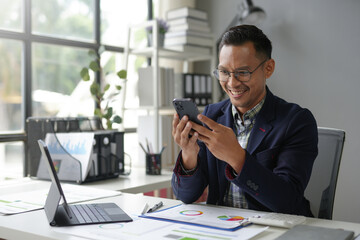 Wall Mural - Cheerful businessman is checking his smartphone while sitting at his desk in a bright office