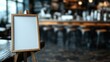 © Dulemegapixel - A blank wooden easel stands prominently in a cozy café with warm blurred lights in the background, creating an inviting and creative ambiance.