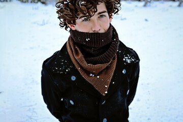 A young man in his twenties with curly hair in a coat and scarf in front of a winter landscape. Selective focus. Snowflakes on the coat and hair.