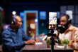 © DC Studio - Selective focus of a smartphone on tripod being used to record two african american bloggers discussing different topics. Black men meeting in home studio using a mobile device to film a show.