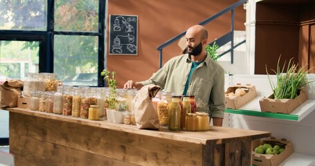 Canvas Print - Smiling client looking to buy spices from bulk products section, zero waste eco store with healthy food alternatives and natural produce. Young buyer choosing bio pantry supplies.