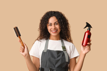 Poster - Beautiful young happy African-American hairdresser with brush and spray on beige background