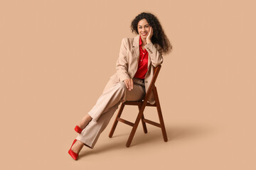 Wall Mural - Beautiful young happy African-American woman in stylish suit sitting on chair against beige background