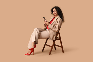 Poster - Beautiful young happy African-American woman in stylish suit with mobile phone sitting on chair against beige background