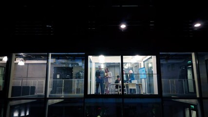 Poster - Businessman and a Female Executive Manager Standing Next to a Window in Office, Using a Laptop Computer. Team of Specialist Having a Meeting, Discussing a Report. View From Outside the Window at Night