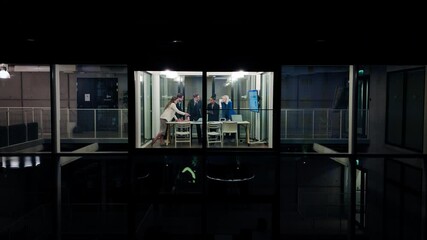Aufkleber - Businessmen and Businesswomen Standing in Office, Talking and Using a Laptop Computer. Team of Specialist Having a Meeting, Discussing Corporate Data. View From Outside the Window at Night