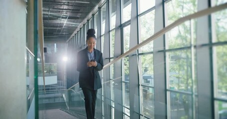 Poster - Black Female Manager Walking in a Modern Glass-Walled Building, Using Her Smartphone on the Move. Bright, Natural Light Highlights the Contemporary Workspace, Happy Young Woman Commuting to Office