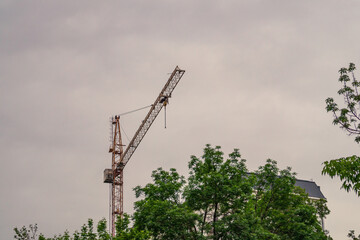 Tower-type construction crane against background of a cloudy sky, erecting a high-rise building located behind large trees. Workplaces associated with working at height, danger of dizziness and panic.