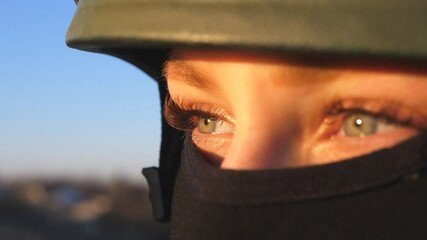 Wall Mural - Close up green eyes of female ukrainian army soldier looking at sunset. Sight of young military woman in helmet and balaclava at countryside. Invasion of territory Ukraine. End of war in Europe