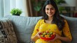 © The Little Hut - Happy indian pregnant woman eating fruits salad while sitting on sofa at home  concept of healthy lifestyles Maternal Pregnancy nutrition : Generative AI