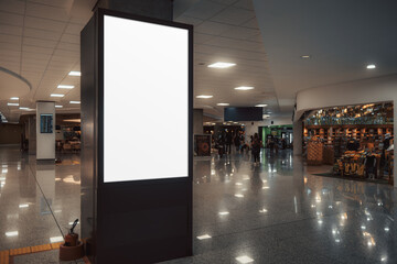 A low-key shot of a vertical blank billboard mock-up stands prominently in the middle of a bustling airport terminal or a shopping mall, with shops in the background, ready for custom advertisements