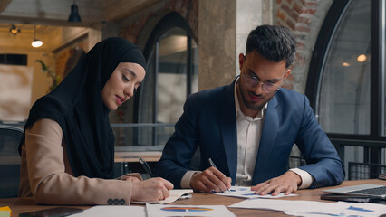 Two business colleagues co-workers talk discuss corporate work at office cafe multiracial male female employees Indian man muslim woman in hijab diverse businesswoman businessman write tasks laptop