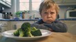 © VK Studio - A young boy with a comically exaggerated frown sits at a table, reluctant to eat a plate of broccoli in front of him.