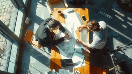 Wall Mural - Two men sit at a large wooden table in a sunlit office, deeply engaged in a discussion over blueprints and documents, with a bird's eye view capturing their collaboration.