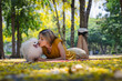 © kamonrat - Woman Lying on Grass, Kissing Her Happy Fluffy Dog in a Sunlit Park, Surrounded by Trees and Yellow Leaves