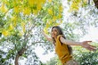 © kamonrat - Joyful Woman in a Sunlit Park with Arms Outstretched, Surrounded by Lush Greenery and Yellow Blossoms, Embracing the Beauty of Nature