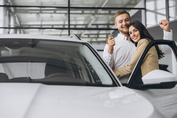 Happy young lovely couple in casual wear hugging while buying first new family car together in dealership. Man shows car keys in showroom.