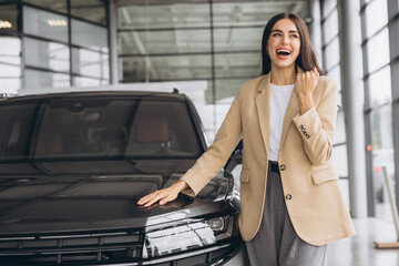 Wall Mural - Charming young Caucasian woman hugging hood of new car and show fist and say yes in modern bright car dealership. Happy girl buyer rejoices after making her dream come true.