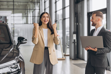 Wall Mural - Charming young Caucasian woman holding keys of new car and show fist and say yes in modern car dealership. Happy girl buyer rejoices after making her dream come true.