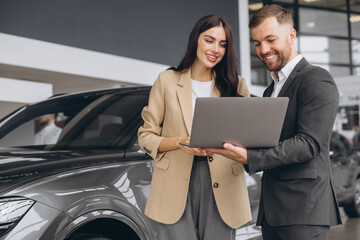 Wall Mural - Male car salesman, recommends a car to a woman buyer, inspects cars in the showroom, the seller uses a laptop to view technical information on the car