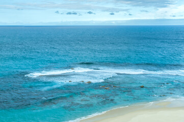 A serene view of the Indian Ocean off the coast of Western Australia. The clear blue water gently meets the sandy shore, creating a tranquil and picturesque seascape perfect for relaxation and escape.