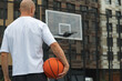 © svetlana_cherruty - Sporty man holding orange basketball ball on sports court. Basketball Hoop. View from back. Urban city park outside.